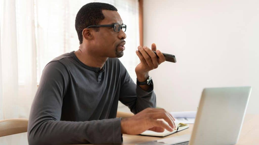 A man using voice commands on a smartphone while working on a laptop.