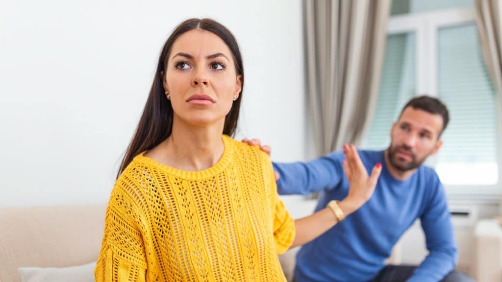 Woman in a yellow sweater holding up her hand to ward off a man in a blue sweater who has his hand on her shoulder.