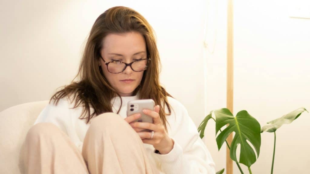 A woman wearing glasses sits in a chair while looking down at a mobile phone screen.
