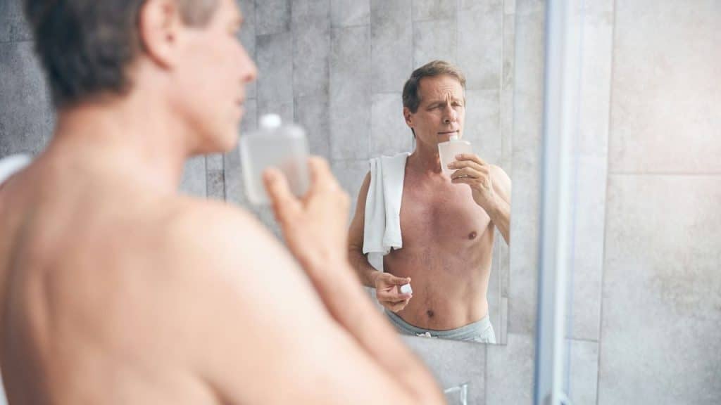 A shirtless man standing in front of a bathroom mirror while smelling a glass bottle.