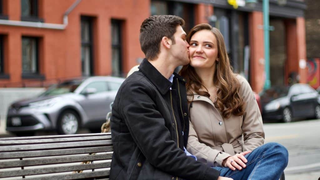 A man kisses a woman’s cheek while they sit together on a park bench outdoors.