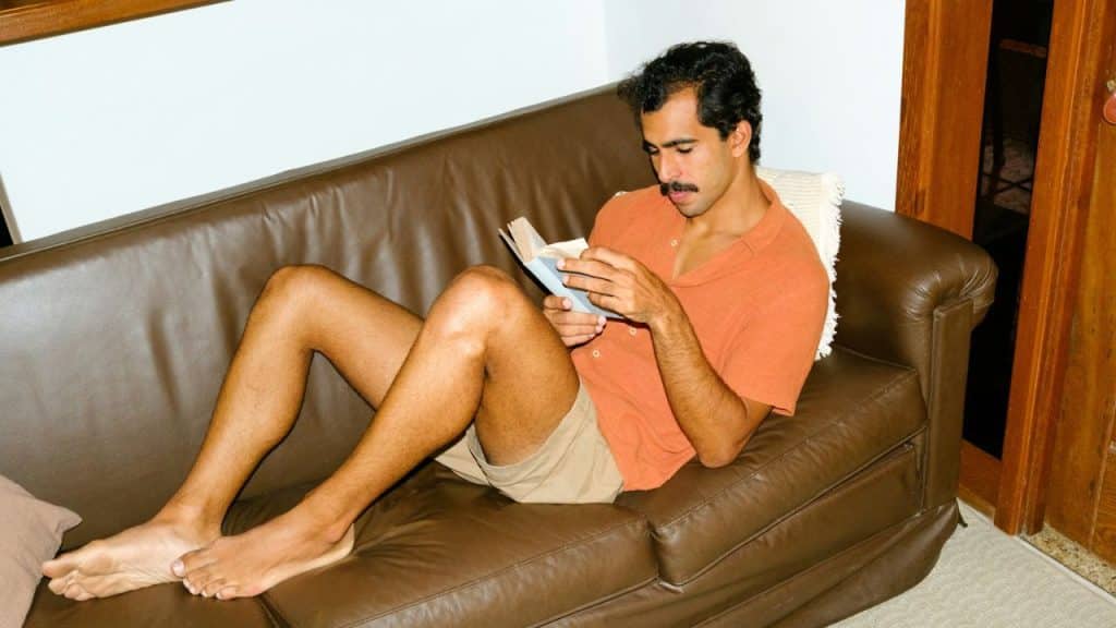 A man with a mustache reclining on a brown leather couch reads a book.