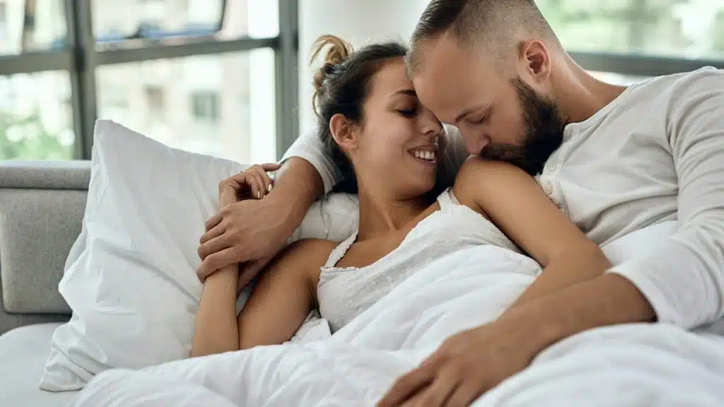 A man and woman lie in bed together under white covers, smiling and holding hands.