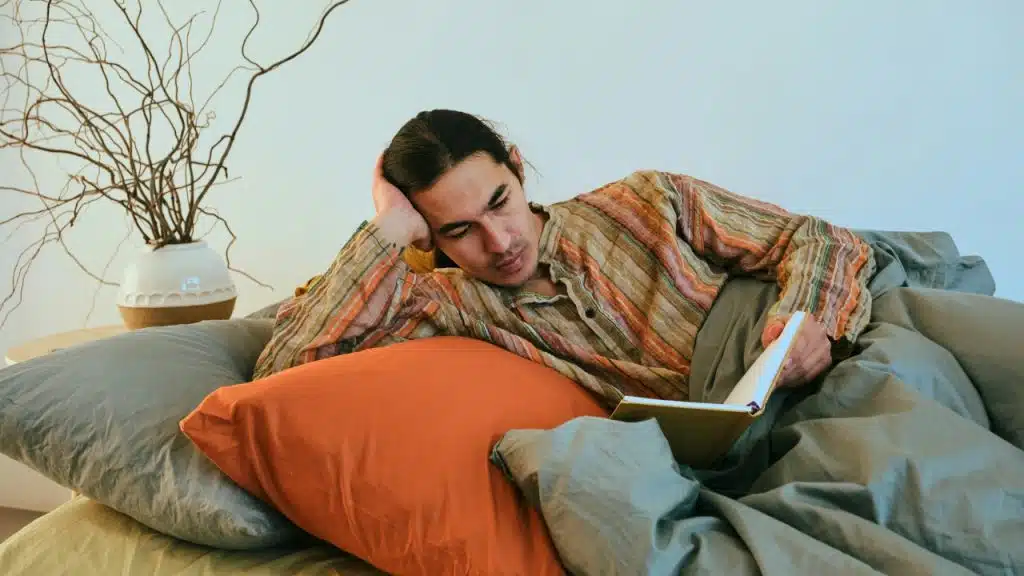 A man in a striped shirt lies in bed and reads a book with one hand.