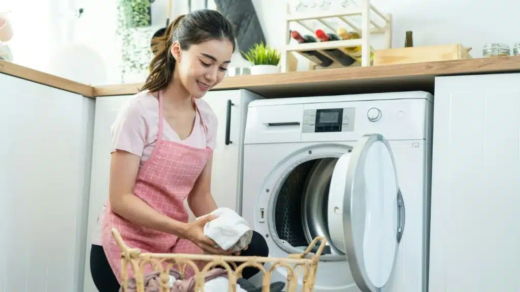 A smiling woman in a pink apron puts white laundry into a front-loading washing machine.