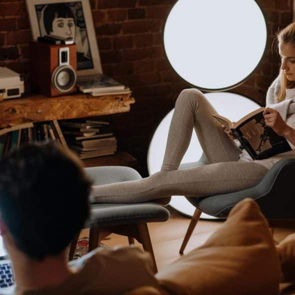A woman reads in a chair while a man uses a laptop in a cozy room.