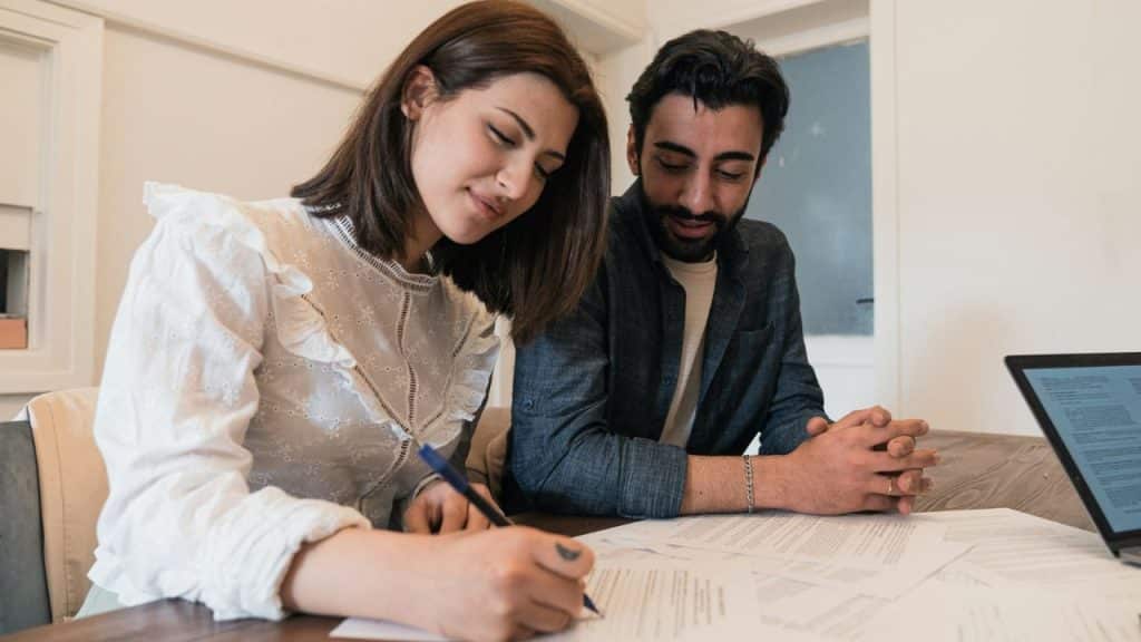 A woman signs a paper document with a pen while a man sits beside her watching.