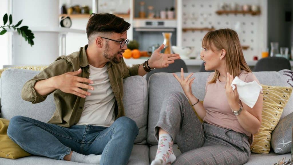 A man and woman sit on a couch gesturing at each other while talking in a kitchen.