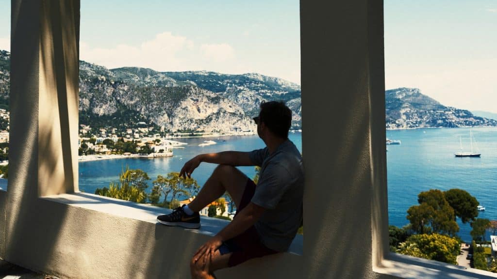 A person sits on a ledge overlooking a blue bay surrounded by green coastal mountains.