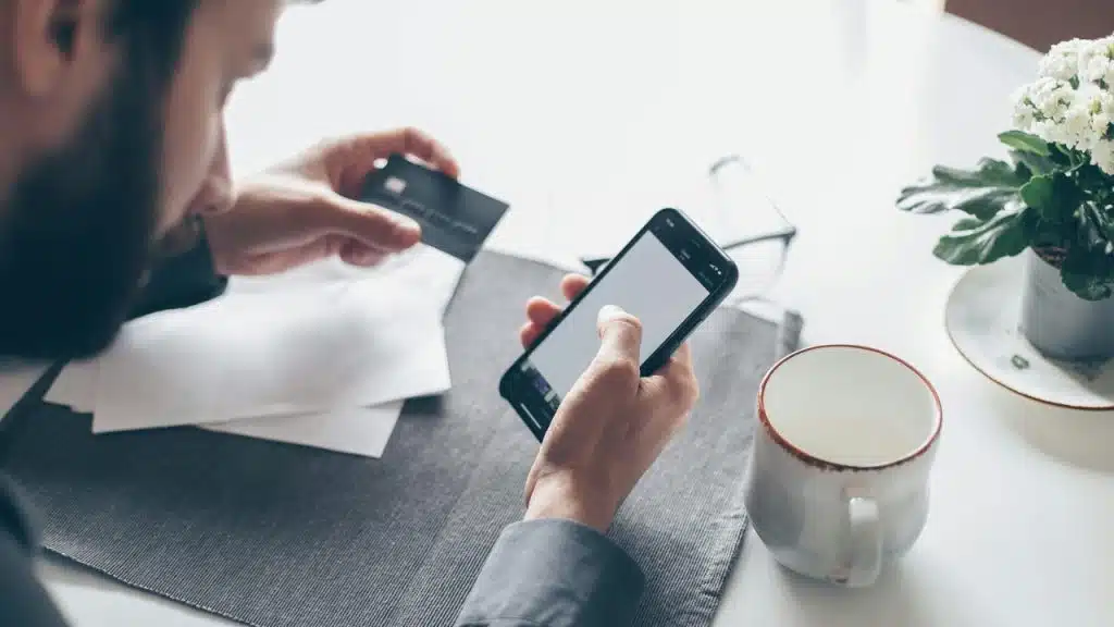 A man holds a black card and a smartphone over a white table with a mug.