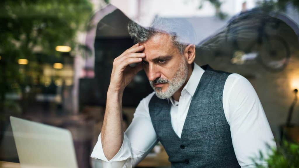 A man with grey hair rests his head on his hand while looking at a laptop.