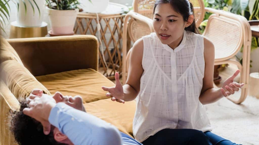 A woman gestures with both hands while speaking to a man lying on a sofa.