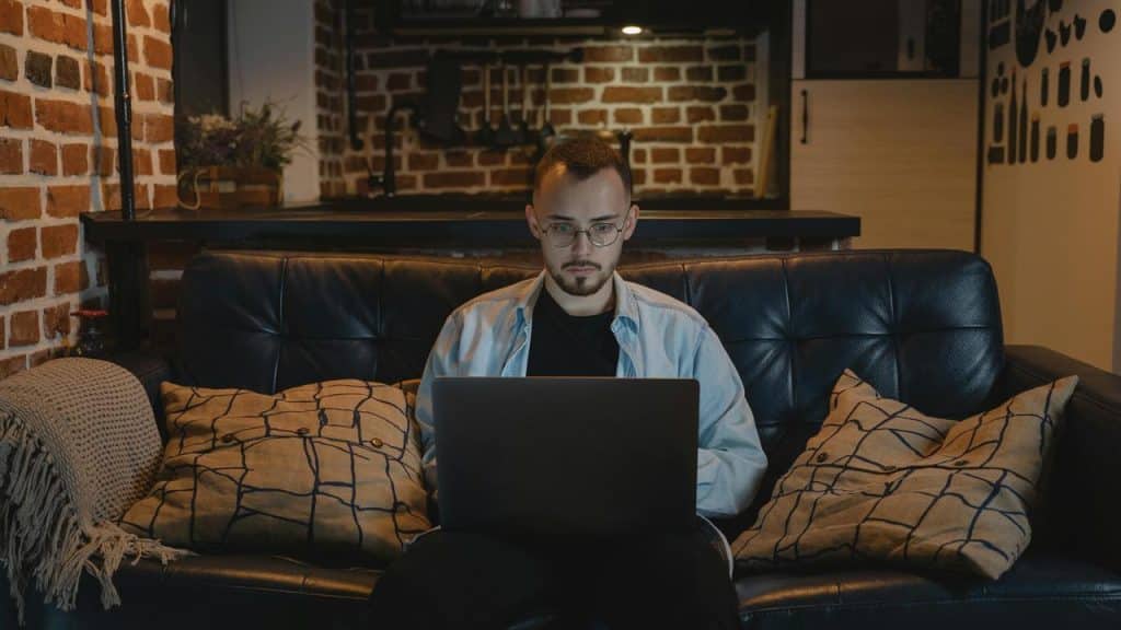 A man with glasses works on a laptop while sitting on a couch at home.