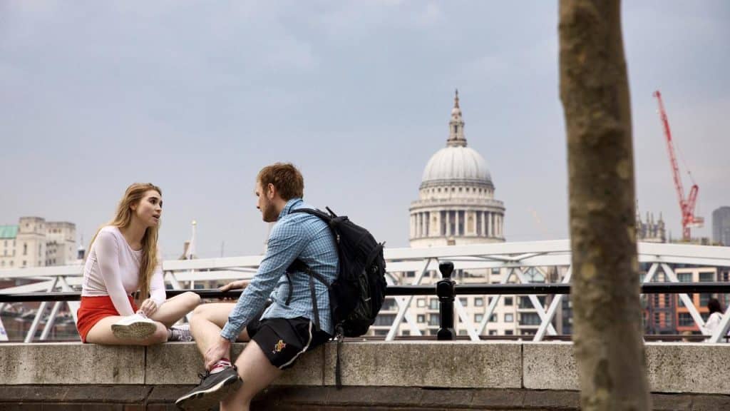 A man and woman sitting by a riverside railing with a city dome in the background.