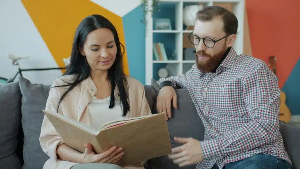 A woman and a man sit on a couch together while looking at a book.