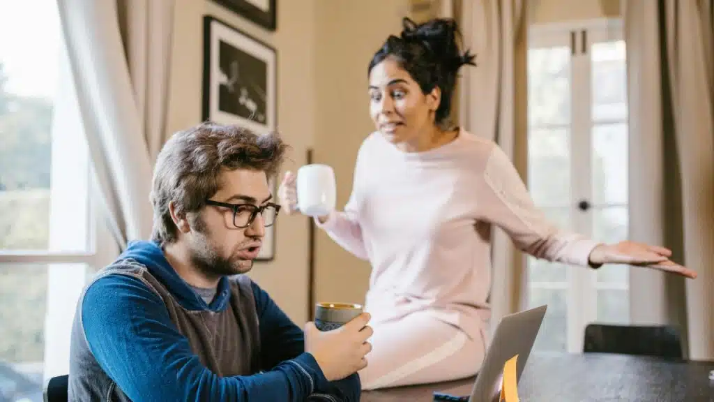 A woman gestures broadly and speaks toward a man sitting at a table with a laptop.
