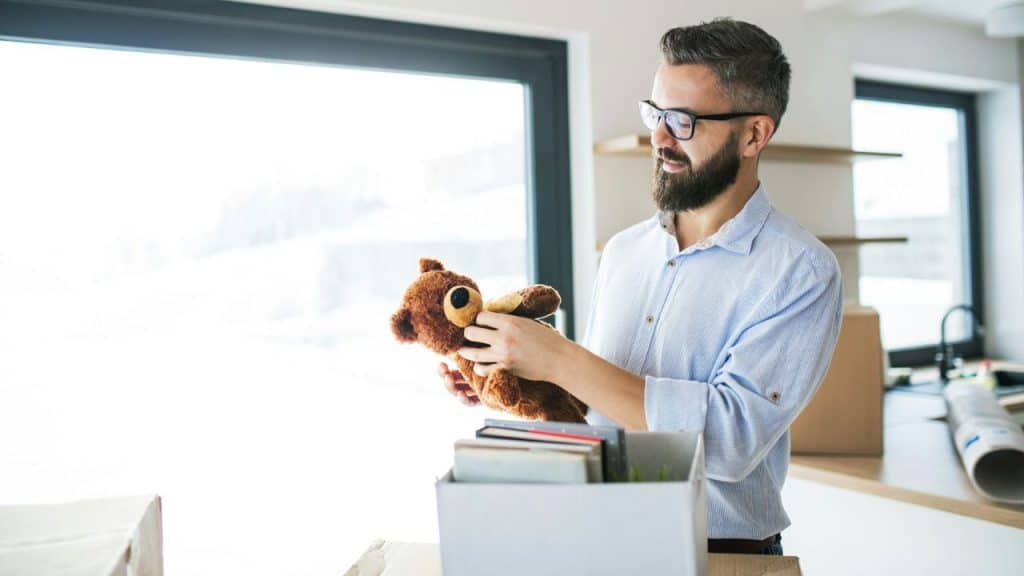 A man with a beard and glasses holds a brown teddy bear near a cardboard box.