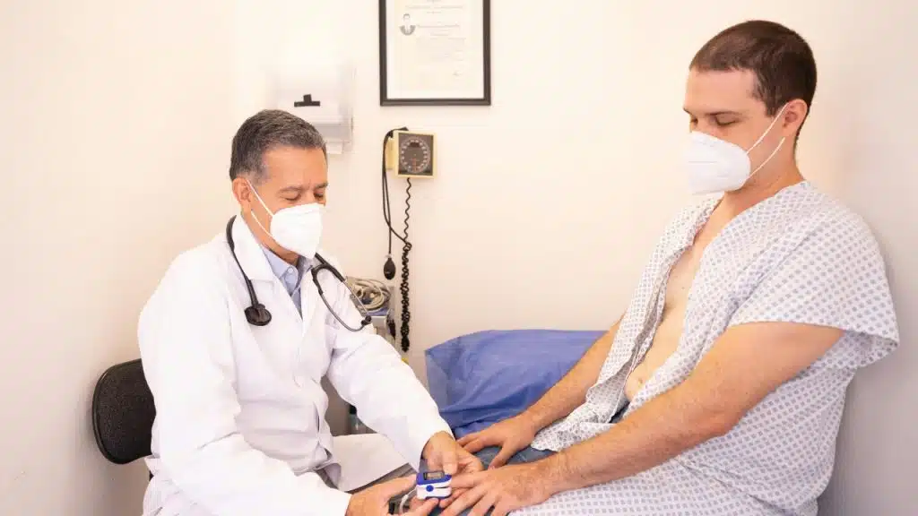 A doctor in a white coat and mask uses a finger pulse oximeter on a patient.