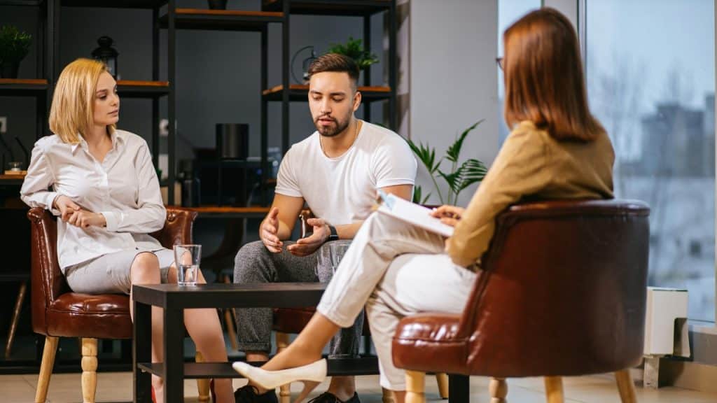 A man and woman sit on a sofa talking to another person holding a clipboard.