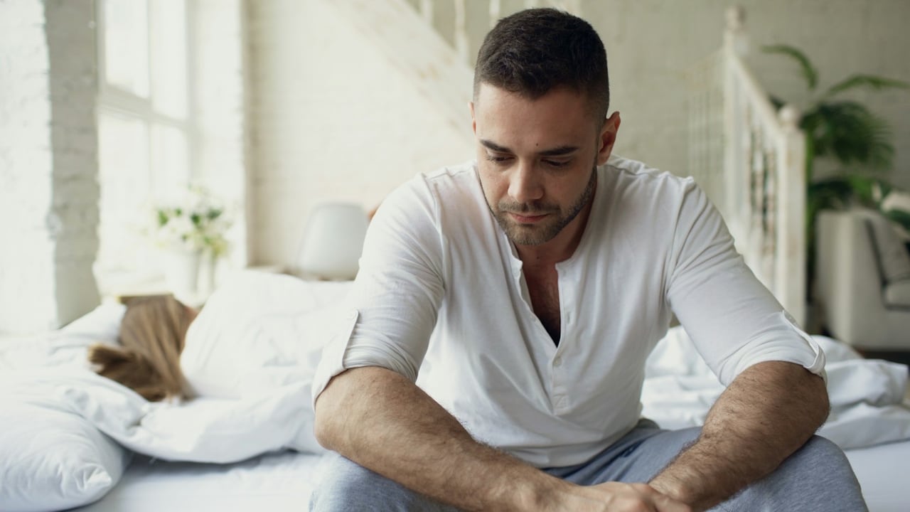 A sad man sitting on the edge of the bed looking down while a woman lies asleep or turned away behind him.