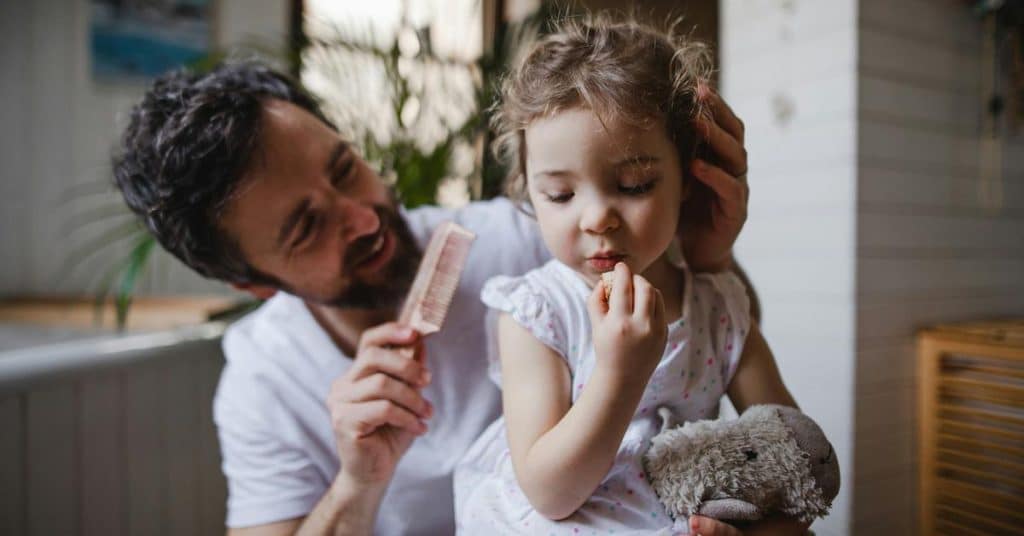 A father gently brushing a young child’s hair while she holds a stuffed toy.
