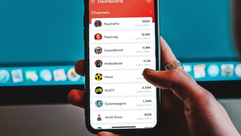 A hand holds a smartphone showing a list of social media channels over a computer monitor.