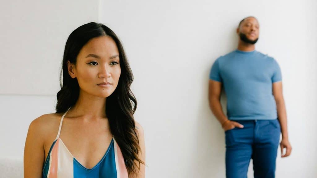 A woman standing in the forefront while a bearded man leans against a wall in the background.