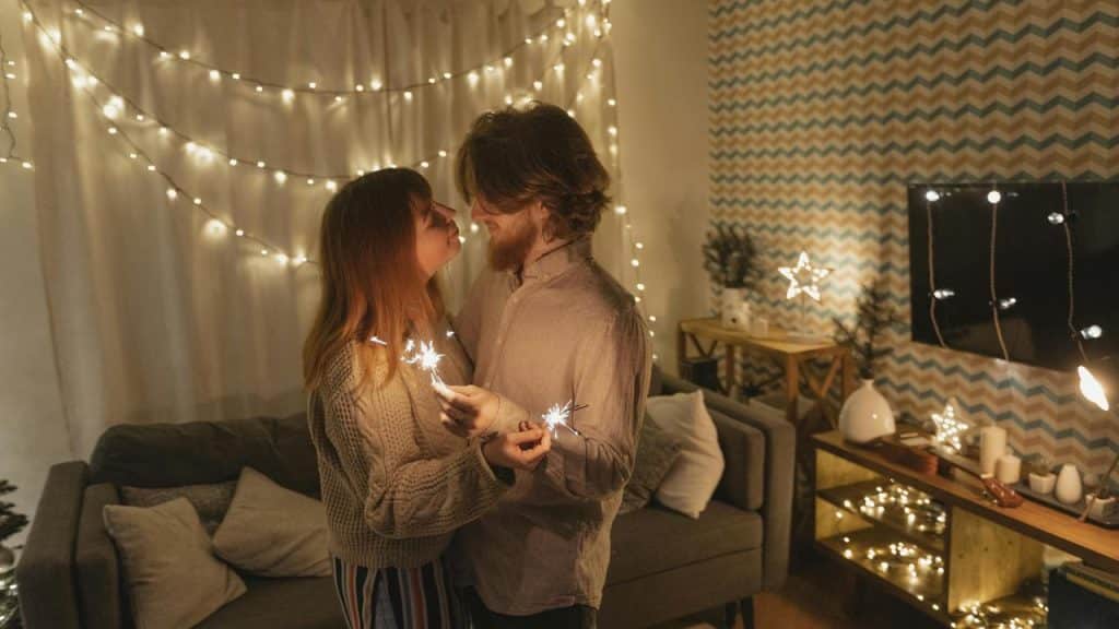 A couple holding sparklers and standing close together in a warmly lit, festive room.