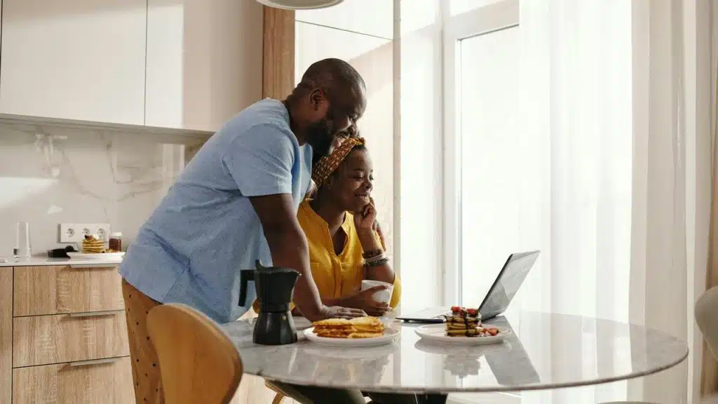 A couple smiling while looking at a laptop together at a kitchen table.