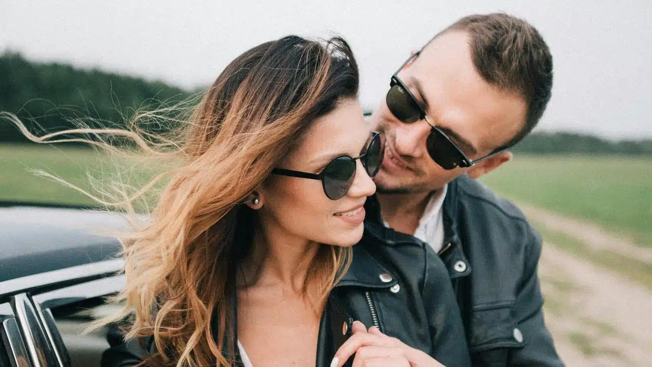A couple wearing sunglasses smiling at each other beside a car in the countryside.