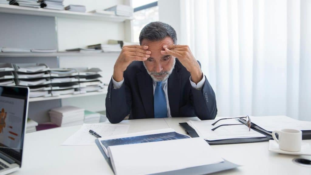 A man in a suit sitting at a desk holding his head in his hands.