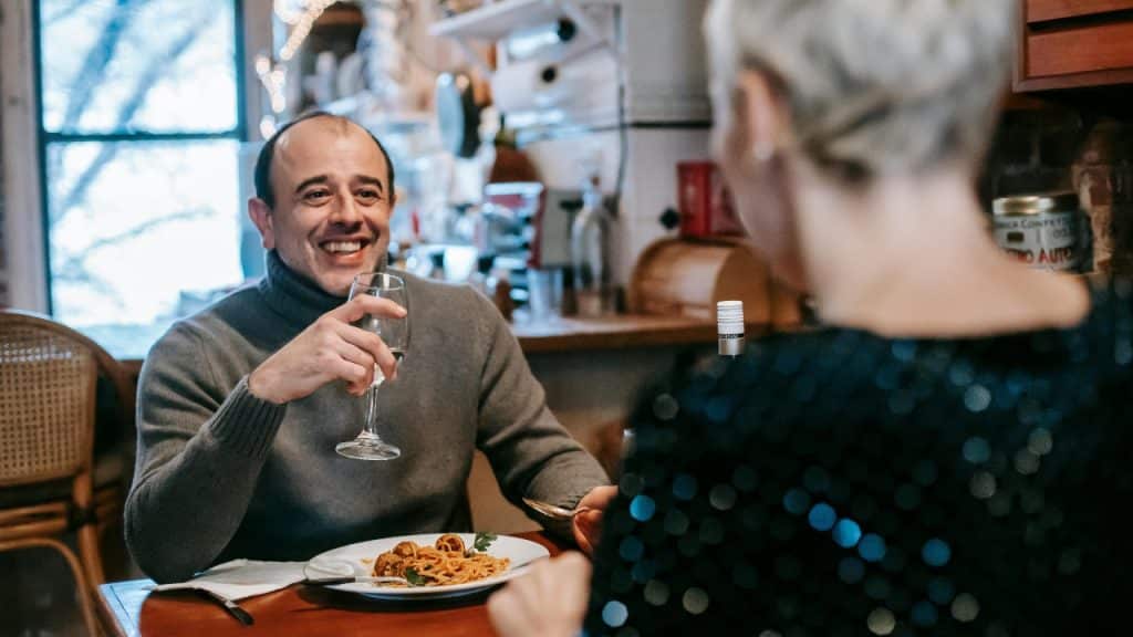 A smiling man holds a wine glass at a dinner table while facing another person's back.