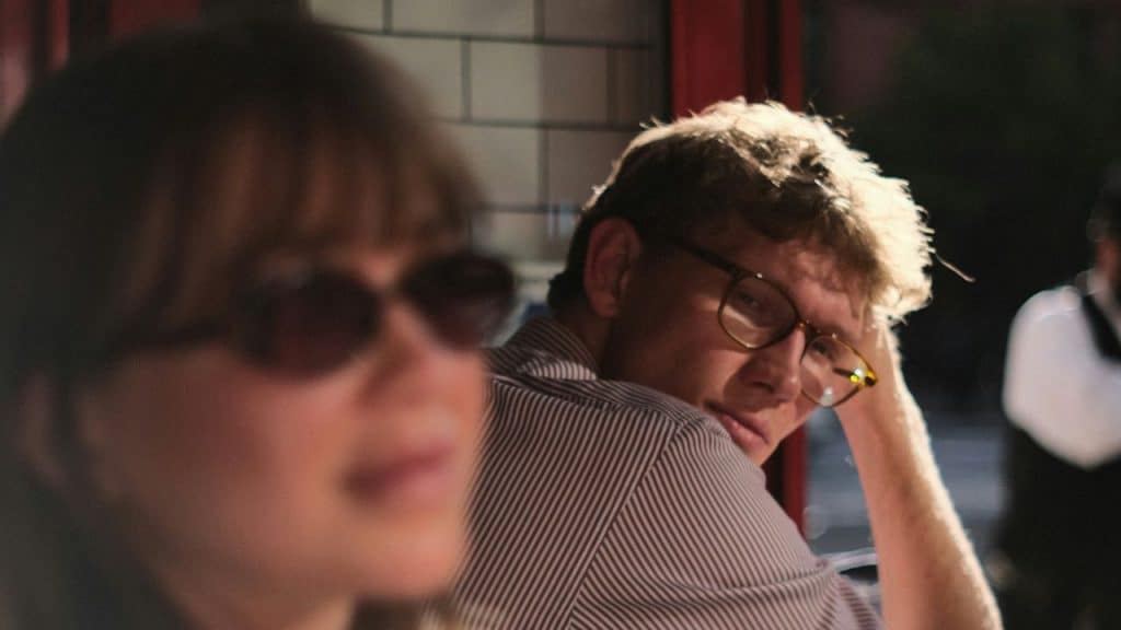 A man wearing glasses looks over his shoulder toward a woman wearing sunglasses in foreground.