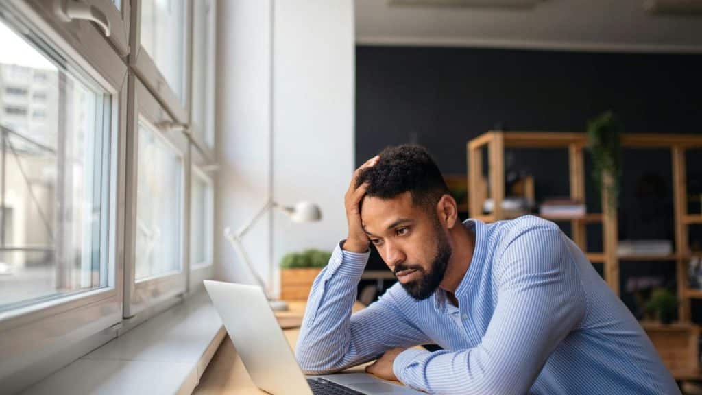 A man sits at a desk with his hand on his head, looking at a laptop.