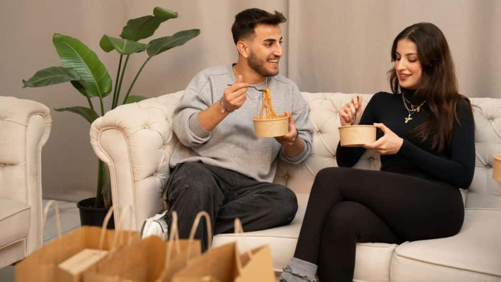 A man and woman sit on a white sofa eating noodles from takeout containers.