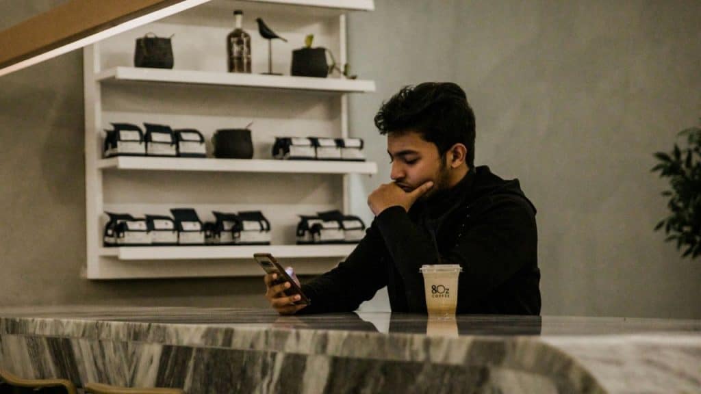 A man sits at a marble counter looking at a smartphone next to a plastic cup.