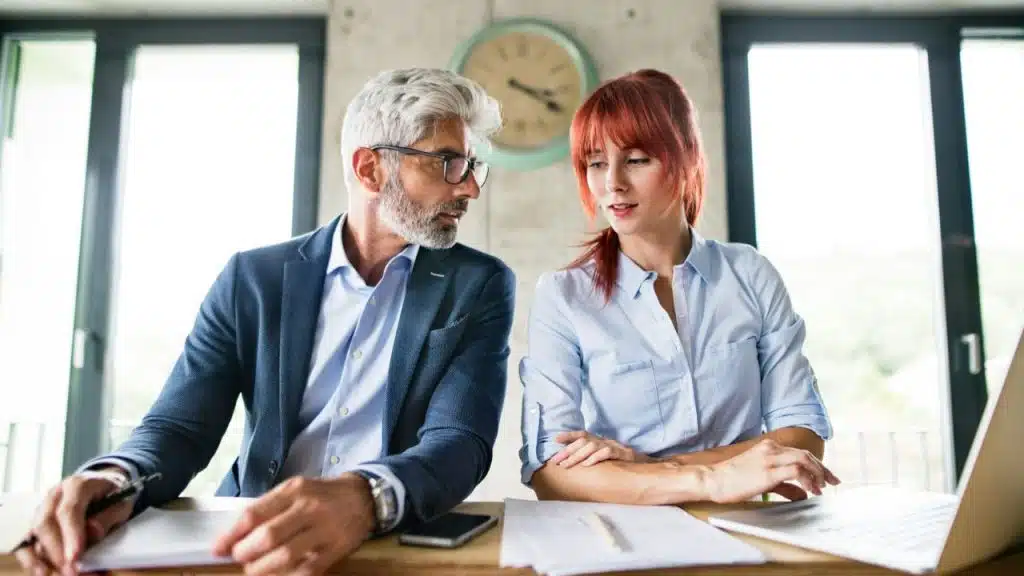 A man with grey hair and glasses looks at a woman with red hair.