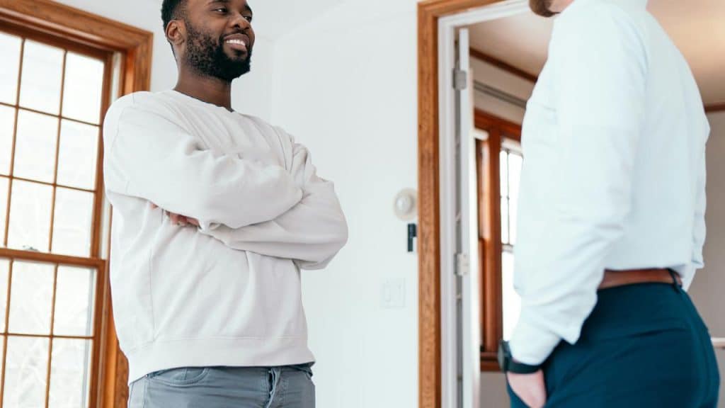 A man smiling with his arms crossed while talking to another man indoors.