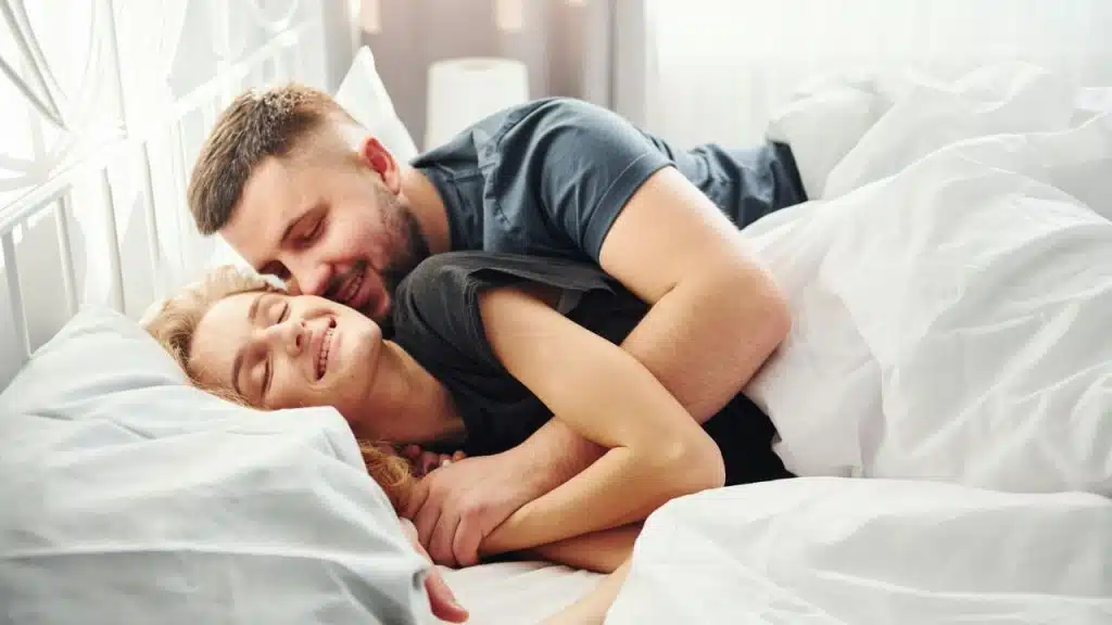 A smiling man and woman with closed eyes embrace while lying in a white bed.