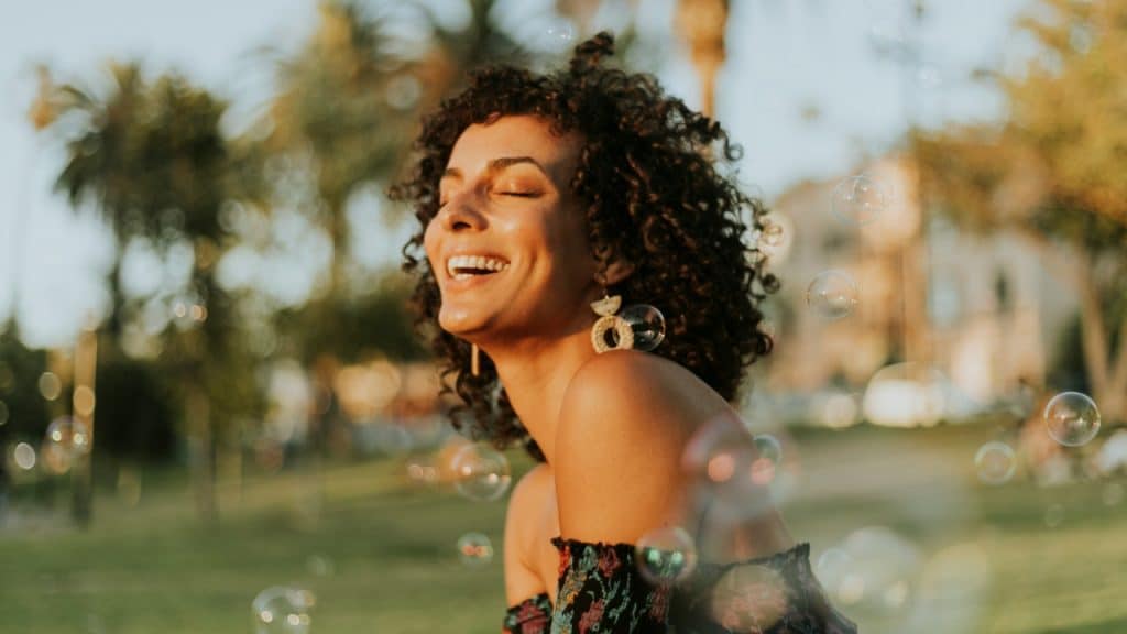 A laughing woman with curly hair stands outdoors surrounded by floating bubbles in sunlight.