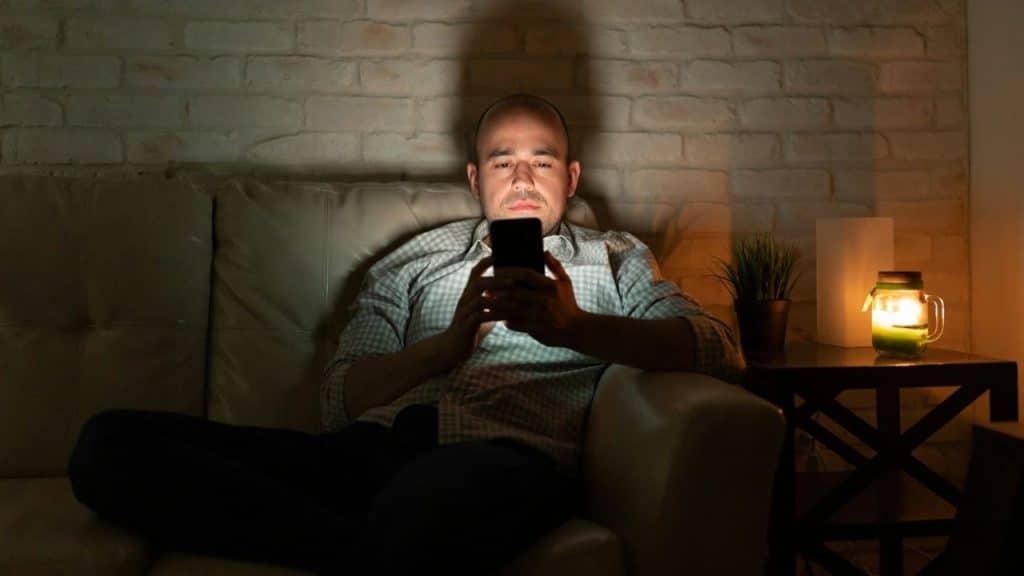 A man sits alone on a sofa in a dark room looking at a glowing smartphone.