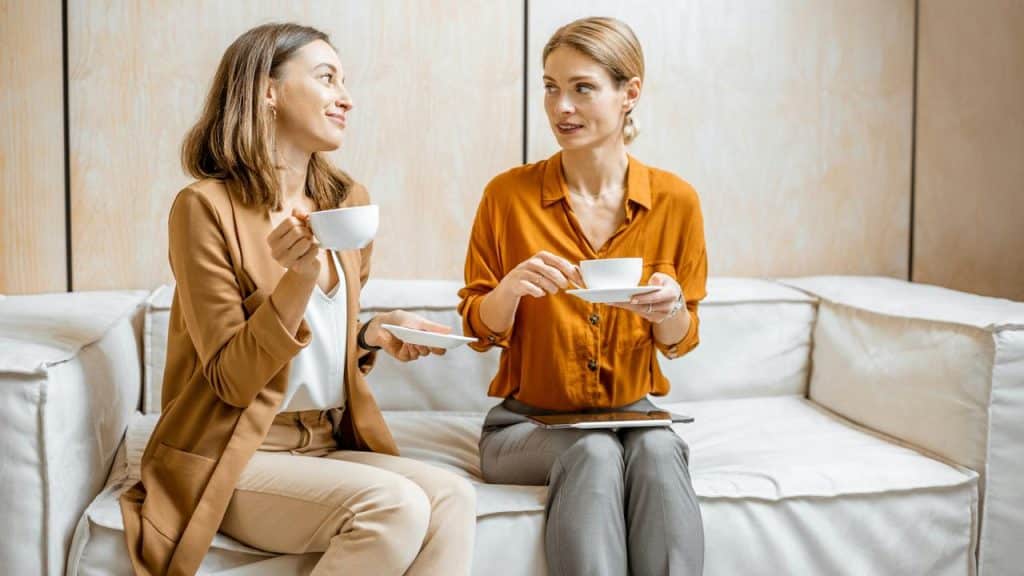 A pair of women sitting on a couch, chatting while holding coffee cups.