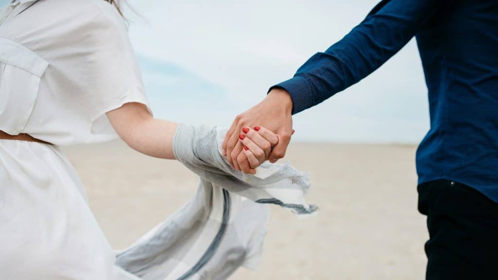 A medium shot shows a woman in a white dress leading a man in a blue button-down shirt by the hand across a sandy, open landscape.