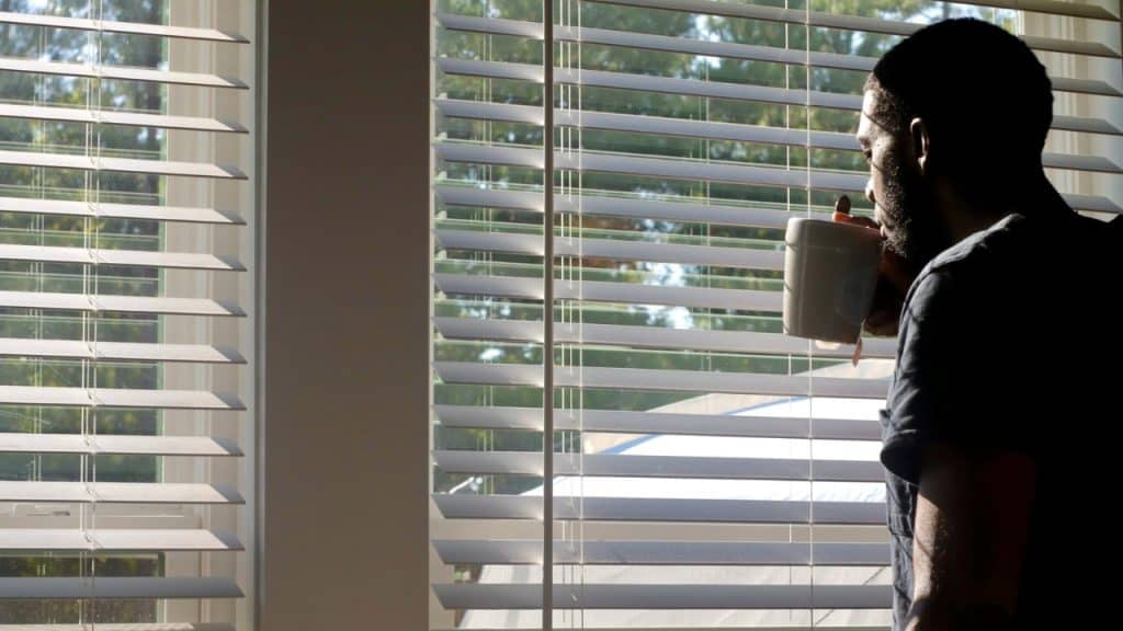 A man in silhouette drinks from a white mug while looking through closed window blinds.
