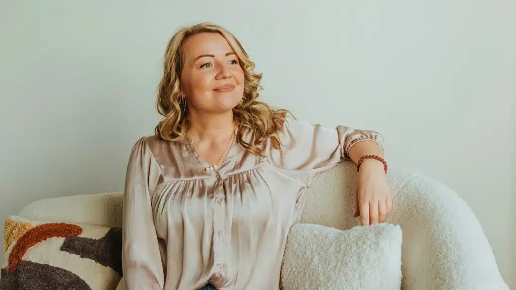 A smiling woman sitting on a couch and resting her arm on it.