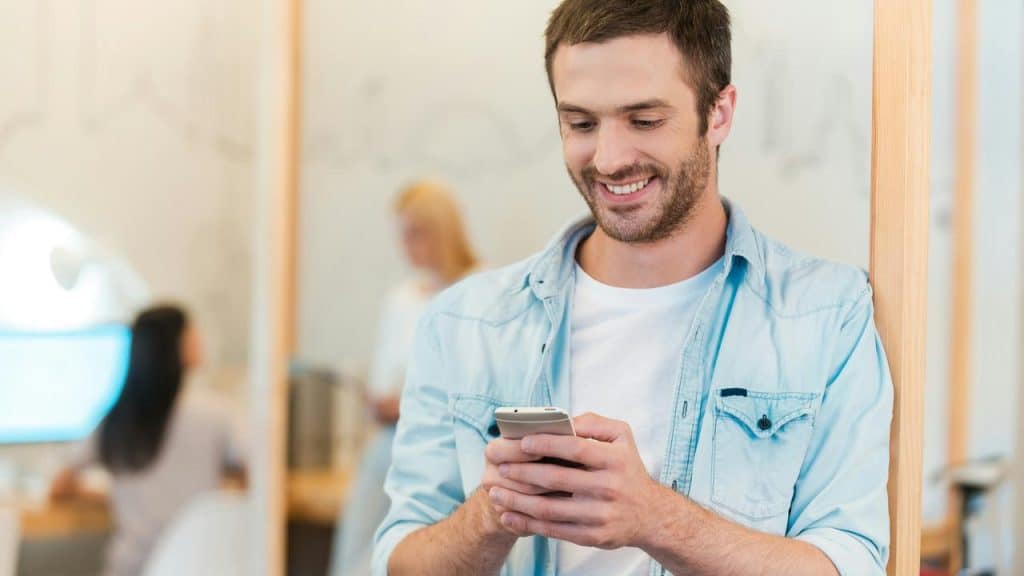 A smiling man looks at his phone while standing indoors.