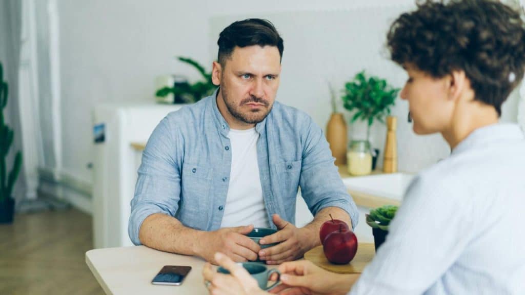 A man with a furrowed brow sits at a table looking at a blurred person.