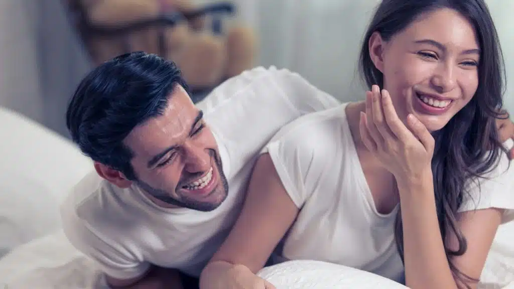 A man and woman lie on a white bed, laughing together with their eyes closed.