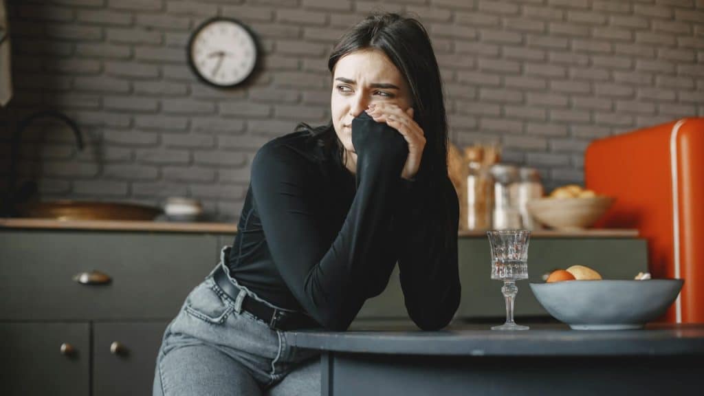 A woman in black sits at a table with her hands clasped, looking to the side.
