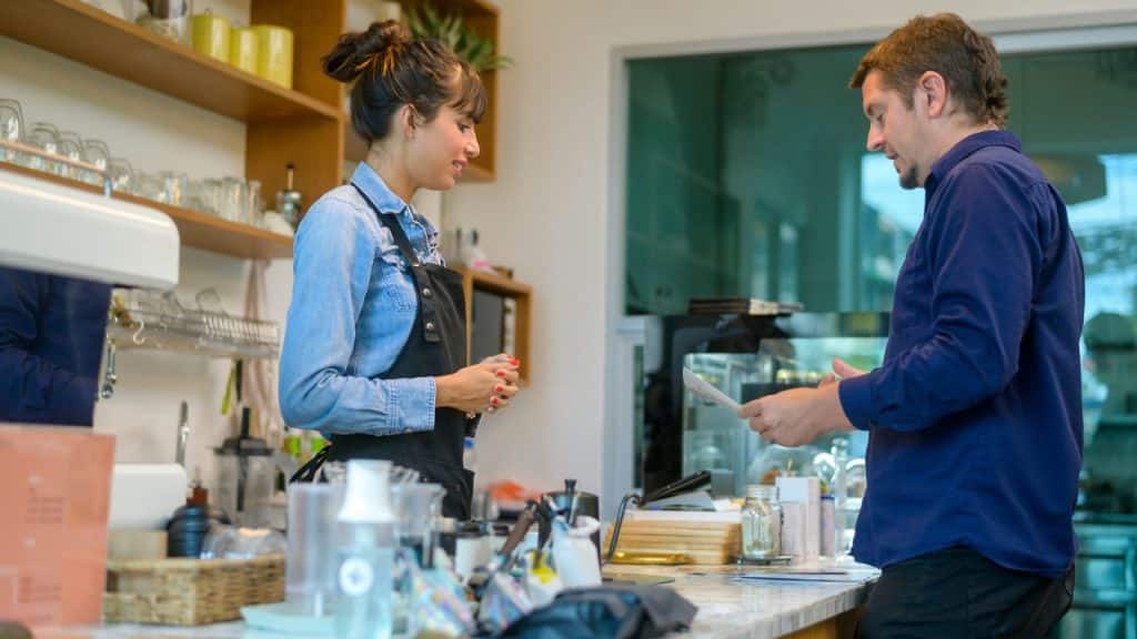 A woman in a cafe apron stands behind a counter while a man reviews a paper.